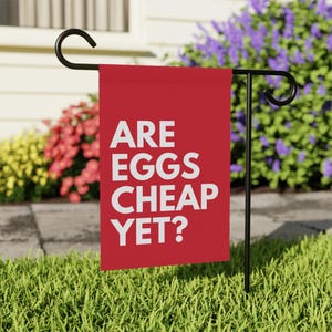May include: A red garden flag with white text that reads "Are eggs cheap yet?" The flag is hanging from a black metal stand in a grassy yard.