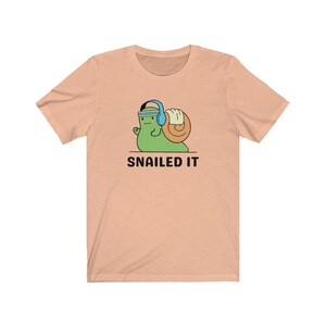 K&ouml;nnte beinhalten: Ein pfirsichfarbenes T-Shirt mit einer Cartoon-Schnecke, die Kopfh&ouml;rer tr&auml;gt und ein Gewicht hebt. Der Text "SNAILED IT" ist unter der Schnecke aufgedruckt.