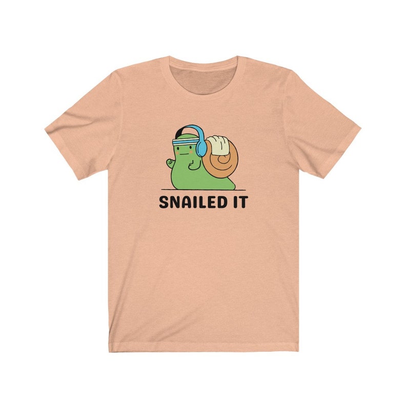 K&ouml;nnte beinhalten: Ein pfirsichfarbenes T-Shirt mit einer Cartoon-Schnecke, die Kopfh&ouml;rer tr&auml;gt und ein Gewicht hebt. Der Text "SNAILED IT" ist unter der Schnecke aufgedruckt.