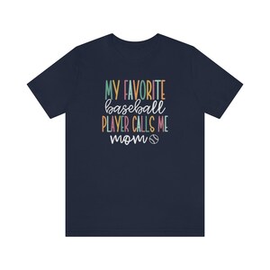 Op de afbeelding: Marineblauw T-shirt met de tekst "MY FAVORITE baseball PLAYER CALLS ME mom" in kleurrijke letters. Een honkbalafbeelding is inbegrepen. Het shirt heeft een ronde hals en is gemaakt van een zacht materiaal.