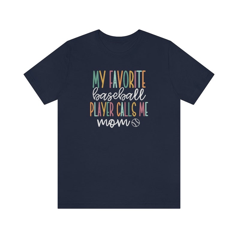 Op de afbeelding: Marineblauw T-shirt met de tekst "MY FAVORITE baseball PLAYER CALLS ME mom" in kleurrijke letters. Een honkbalafbeelding is inbegrepen. Het shirt heeft een ronde hals en is gemaakt van een zacht materiaal.