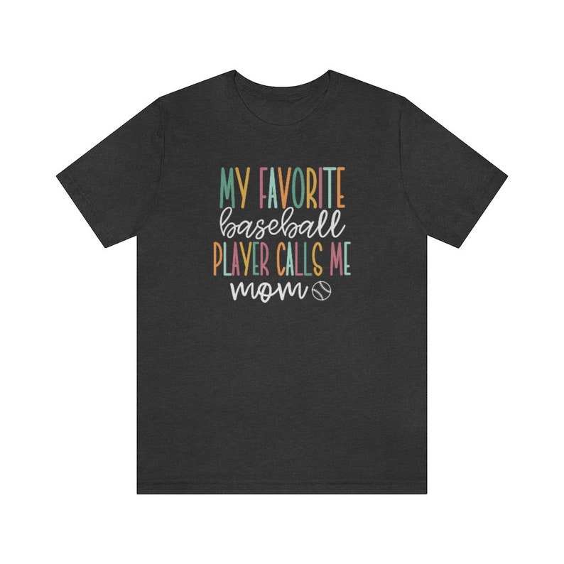 Op de afbeelding: Donkergrijs t-shirt met de tekst "MY FAVORITE baseball PLAYER CALLS ME mom" in kleurrijke letters. Een honkbalafbeelding is opgenomen onder het woord "mom". Het shirt is gemaakt van een zacht materiaal.
