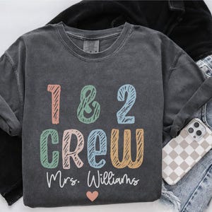 May include: Dark gray crew neck sweatshirt with the text "1 & 2 CREW" in colorful letters. The name "Mrs. Williams" is below the text, with a small pink heart. The sweatshirt is paired with jeans and a phone case.