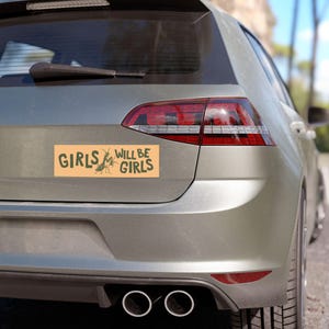 May include: A rectangular bumper sticker with the text "GIRLS WILL BE GIRLS" in dark green, featuring a cartoon insect illustration. The sticker is on the back of a silver car.