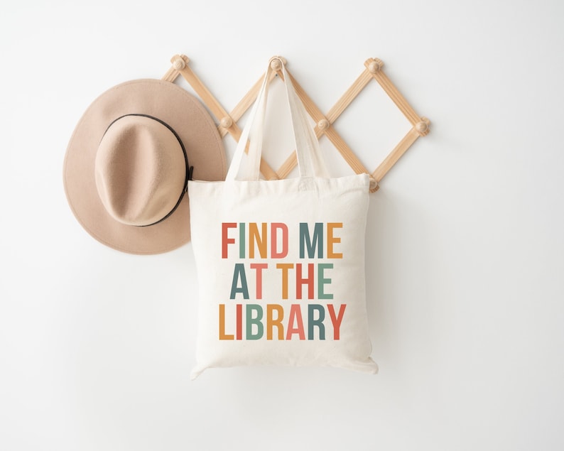 May include: A white canvas tote bag with the text "FIND ME AT THE LIBRARY" in colorful letters. The bag is hanging on a wooden rack with a brown hat hanging next to it.