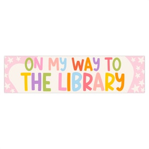 May include: A rectangular sign with the text "ON MY WAY TO THE LIBRARY" in colorful bubble letters. The background is off-white with pink accents and star shapes.