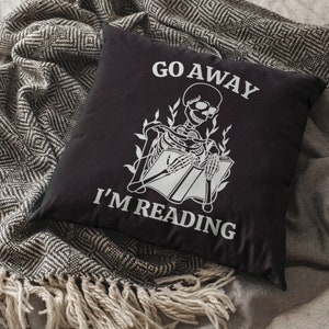 K&ouml;nnte beinhalten: Schwarzes Kissen mit wei&szlig;em Text "Go Away I'm Reading" und einer Skelett-Illustration, die ein Buch liest.