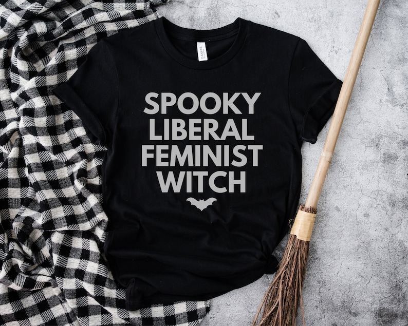 May include: Black t-shirt with the words "SPOOKY LIBERAL FEMINIST WITCH" in white block letters and a bat graphic. A broom and a black and white checkered blanket are in the background. The shirt is made of a soft material.