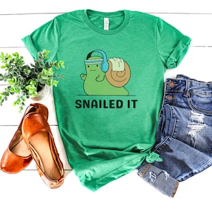 K&ouml;nnte beinhalten: Gr&uuml;nes T-Shirt mit einer Cartoon-Schnecke, die Kopfh&ouml;rer tr&auml;gt und Gewichte hebt. Der Text "SNAILED IT" ist unter der Schnecke gedruckt.