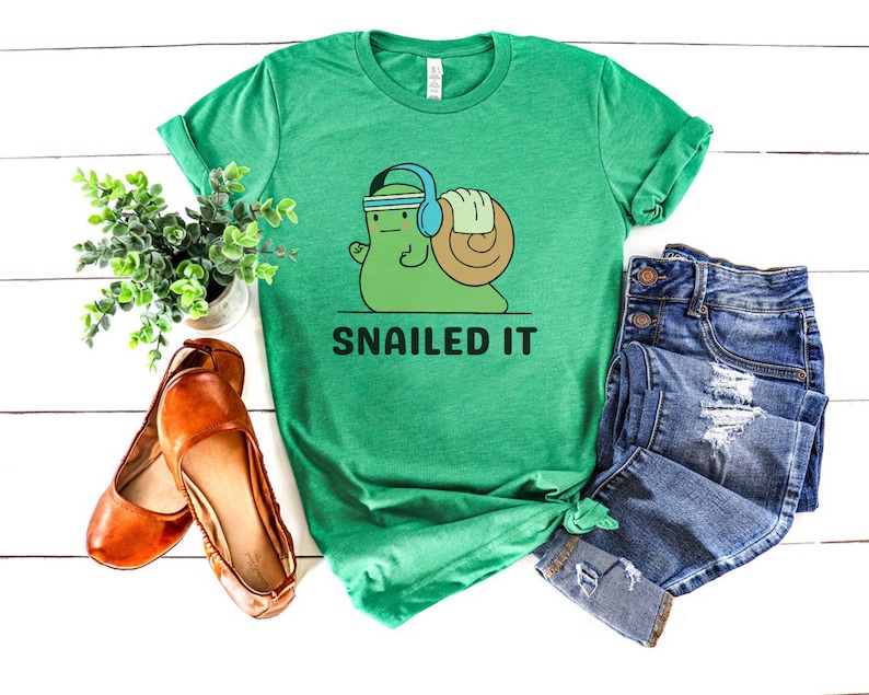 K&ouml;nnte beinhalten: Gr&uuml;nes T-Shirt mit einer Cartoon-Schnecke, die Kopfh&ouml;rer tr&auml;gt und Gewichte hebt. Der Text "SNAILED IT" ist unter der Schnecke gedruckt.