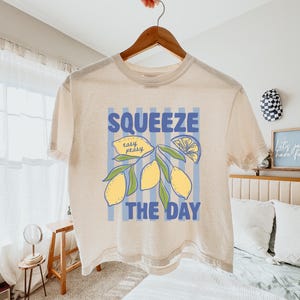 May include: Cream-colored t-shirt with the words "SQUEEZE THE DAY" in blue, along with a lemon illustration. The shirt is hanging on a wooden hanger. The design includes yellow lemons and green leaves.