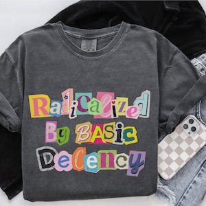 May include: A dark grey sweatshirt featuring the phrase "Radicalized By Basic Decency" in a colourful, cut-out style. The image includes black trousers, light wash jeans, and a phone case with a chequered design.