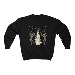 May include: Black sweatshirt with a white Christmas tree and two skeletons wearing Santa hats dancing around it. The background is black with white snowflakes and a crescent moon.
