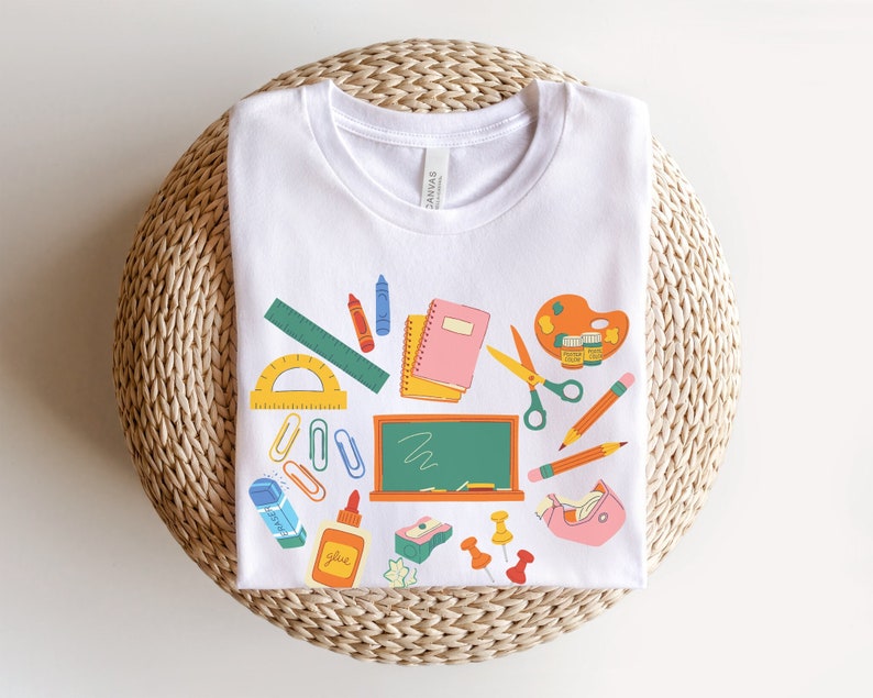 K&ouml;nnte beinhalten: Wei&szlig;es T-Shirt mit einem farbenfrohen Design von Schulmaterialien. Die Grafik umfasst ein Lineal, einen Winkelmesser, Buntstifte, Notizb&uuml;cher, eine Schere, Bleistifte, eine Tafel, Radiergummis, Kleber und Rei&szlig;zwecken. Das Shirt ist aus weichem Material.