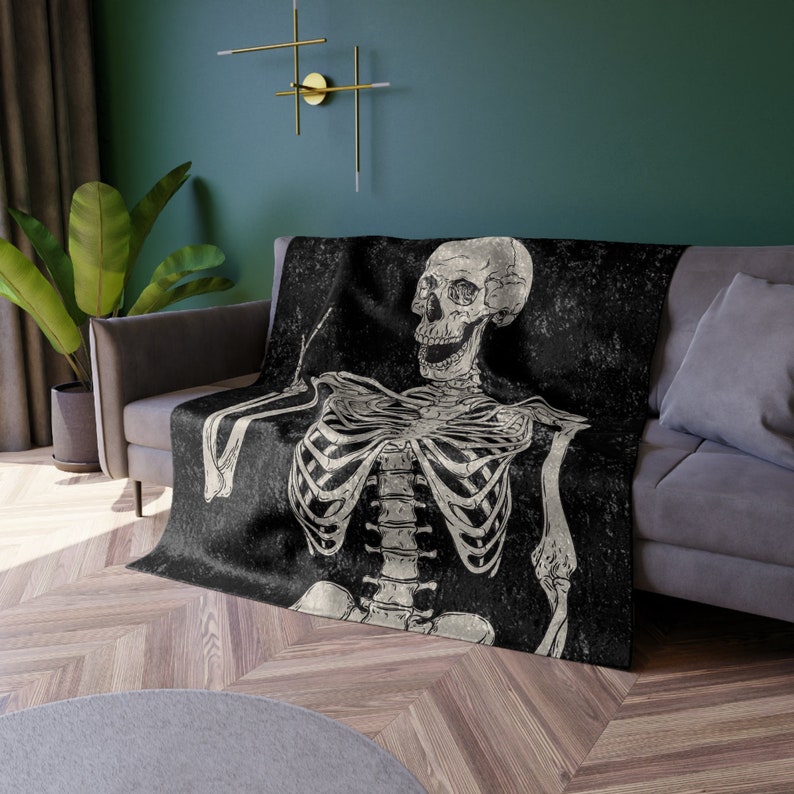 May include: A black and white throw blanket with a skeleton design. The skeleton is sitting with its arms crossed and has a distressed look. The blanket has a vintage, grunge aesthetic.