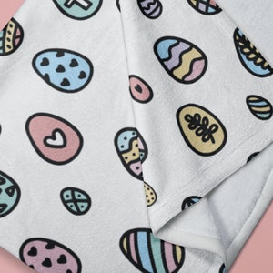 May include: White fleece throw blanket with a repeating pattern of colorful Easter eggs. The eggs are decorated with hearts, leaves, geometric shapes, and stripes.
