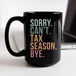 May include: Black ceramic mug with the text "SORRY. CAN'T. TAX SEASON. BYE." in teal, yellow, orange, and pink lettering. The mug has a large handle and is sitting on a white surface.