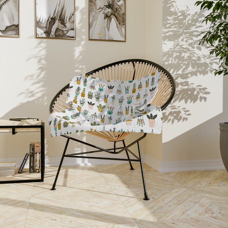May include: A white chair with a black metal frame and a woven rattan seat. The chair is covered with a white throw blanket featuring a pattern of colourful potted plants.