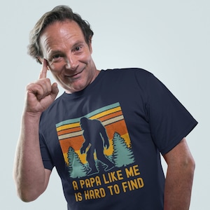 May include: A navy blue t-shirt with a retro-style graphic of a Bigfoot walking through a forest. The text on the shirt reads "A Papa Like Me Is Hard To Find".