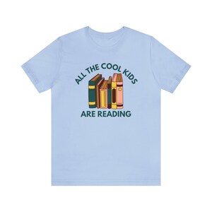 Puede incluir: Camiseta azul claro con el texto "All the cool kids are reading" y una pila de libros coloridos.