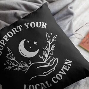 May include: Black throw pillow with white text that reads "Support Your Local Coven". The design also includes a crescent moon, stars, and a hand holding a sprig of leaves.