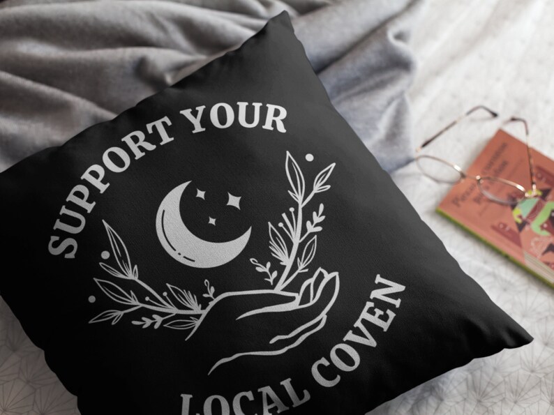 May include: Black throw pillow with white text that reads "Support Your Local Coven". The design also includes a crescent moon, stars, and a hand holding a sprig of leaves.