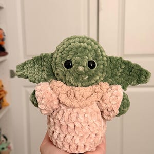 May include: A plush, handmade crochet doll featuring a green head with large black eyes, prominent pointed ears, and a light pink textured robe. The soft, yarn-based creature has a rounded shape and a visible stitch pattern throughout its body.
