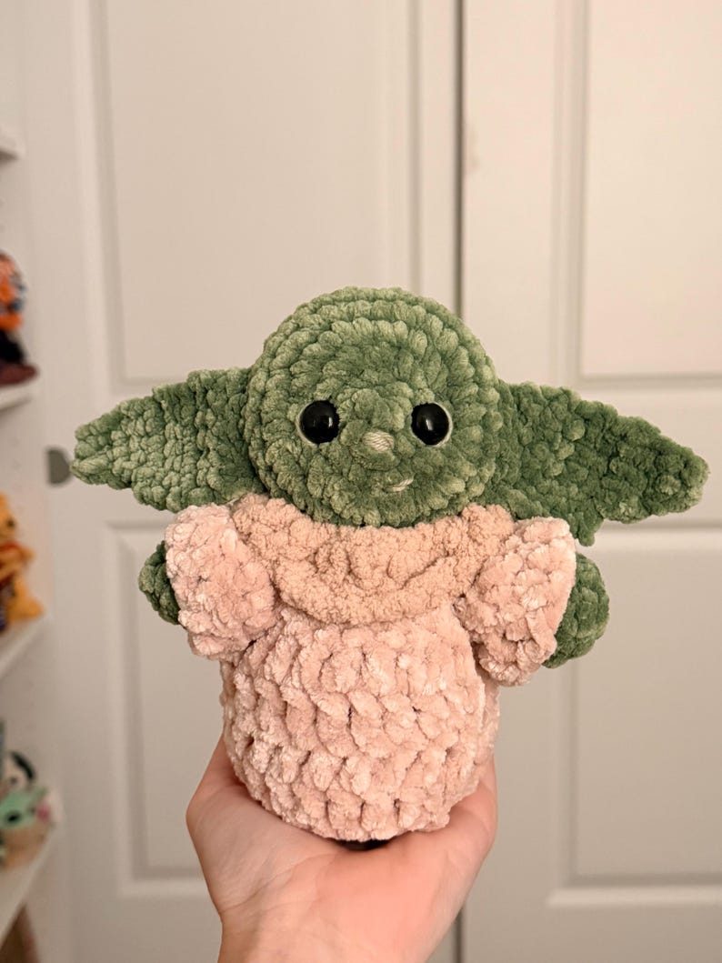 May include: A plush, handmade crochet doll featuring a green head with large black eyes, prominent pointed ears, and a light pink textured robe. The soft, yarn-based creature has a rounded shape and a visible stitch pattern throughout its body.