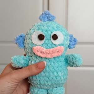 May include: A handmade, plush toy in the shape of a whimsical creature. The toy is primarily light blue with darker blue accents on the head, ears, and arms. It has large, round, brown eyes, a pink mouth, and small feet.