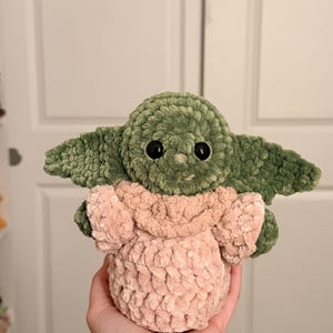 May include: A handmade crochet plush toy featuring a green creature with large pointed ears, black plastic eyes, and a small nose. The figure is wearing a soft, textured light pink robe with a folded collar, crafted from thick, plush yarn.