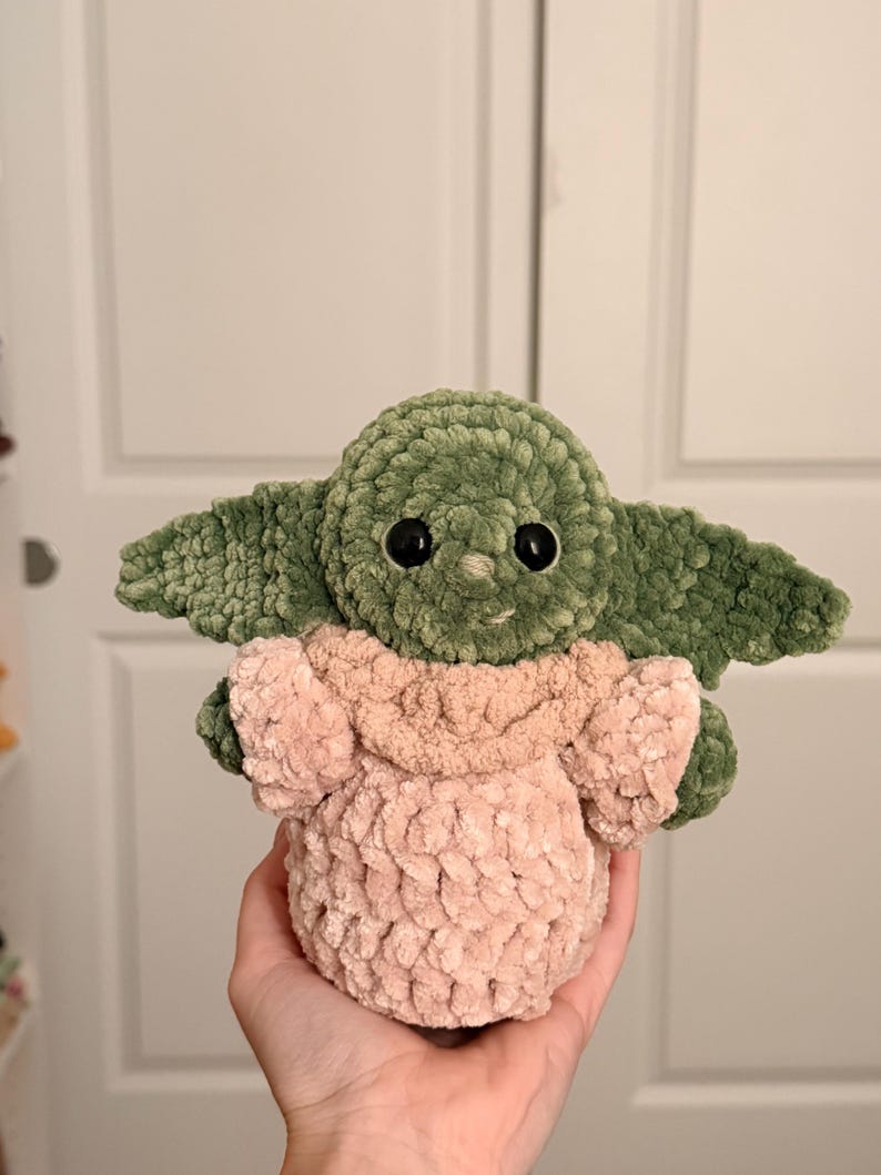 May include: A handmade crochet plush toy featuring a green creature with large pointed ears, black plastic eyes, and a small nose. The figure is wearing a soft, textured light pink robe with a folded collar, crafted from thick, plush yarn.