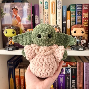 May include: A handmade crochet plush toy featuring a green creature with large ears and black eyes, wearing a light pink textured garment. The figure has a soft, chunky yarn construction with a rounded head and a small, simple facial expression.