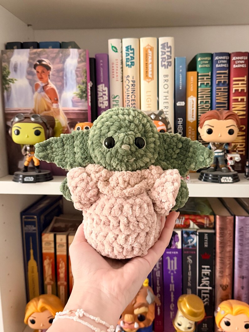 May include: A handmade crochet plush toy featuring a green creature with large ears and black eyes, wearing a light pink textured garment. The figure has a soft, chunky yarn construction with a rounded head and a small, simple facial expression.