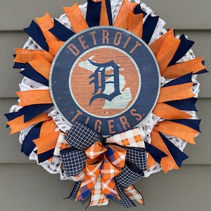 May include: A round wreath featuring the Detroit Tigers logo in the center. The wreath is decorated with orange, navy blue, and white ribbons. A large bow at the bottom adds a decorative touch. The wreath is designed for door or wall decor.