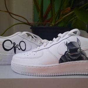 Puede incluir: Zapatillas blancas de cuero con diseños pintados en negro. Un zapato tiene una ilustración en blanco y negro de una criatura con cuernos y un casco. El otro zapato tiene la palabra "Cro" escrita en cursiva.