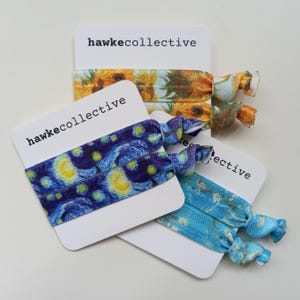 May include: Three sets of hair ties with different printed designs. The first set is yellow and orange with a sunflower pattern. The second set is blue and purple with a starry night pattern. The third set is blue with a watercolor pattern. Each set is packaged on a white card with the text "hawkecollective" printed on it.