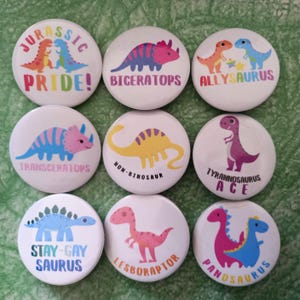 May include: Nine round white buttons with colourful dinosaur illustrations and text. The text includes "Jurassic Pride!", "Bigeratops", "Allysaurus", "Transceratops", "Non-Binosaour", "Tyrannosaurus Ace", "Stay-Gay Saurus", "Lesboraptor", and "Panosaurus".