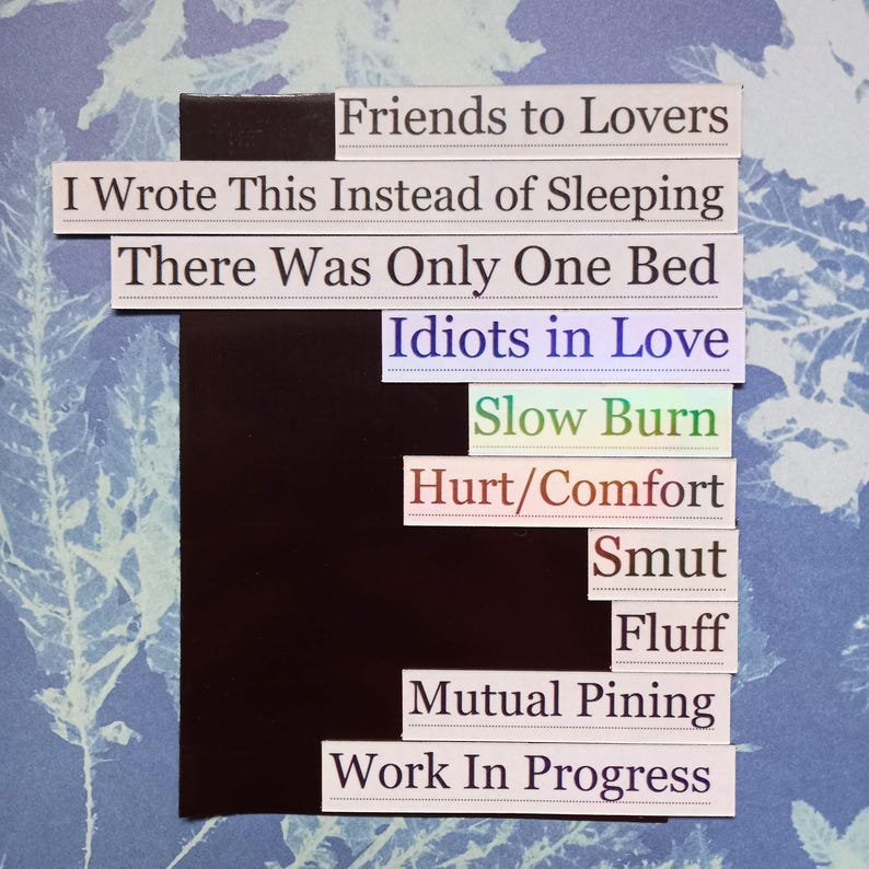Op de afbeelding: Een collage van witte rechthoekige labels met zwarte tekst, gerangschikt op een blauw-witte bloemenachtergrond. De labels bevatten zinnen als "Friends to Lovers", "Idiots in Love" en "Work In Progress".