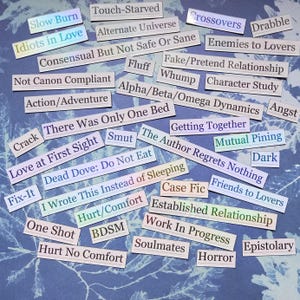 Op de afbeelding: Een verzameling rechthoekige papieren labels met verschillende zinnen, waaronder "Slow Burn", "Idiots in Love" en "Enemies to Lovers". De labels liggen op een blauw, getextureerd oppervlak, sommige met een regenboogglans.
