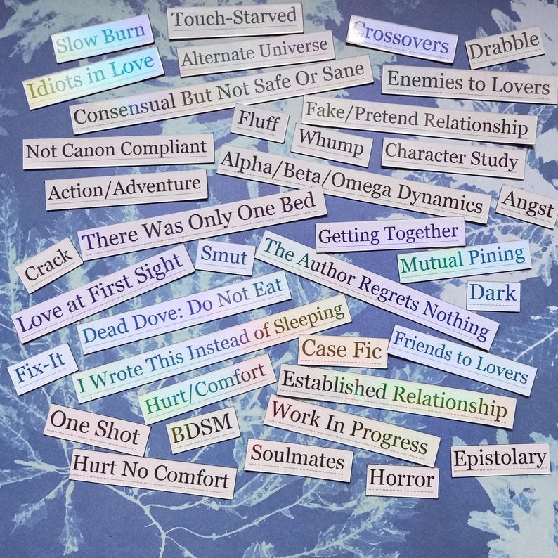 Op de afbeelding: Een verzameling rechthoekige papieren labels met verschillende zinnen, waaronder "Slow Burn", "Idiots in Love" en "Enemies to Lovers". De labels liggen op een blauw, getextureerd oppervlak, sommige met een regenboogglans.