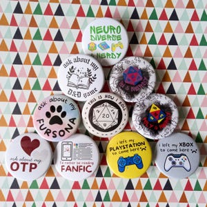 May include: A selection of circular badges with diverse designs and text. Some badges display phrases such as "Ask about my OTP" and "I'd rather be reading FANFIC." Other badges feature gaming themes, including dice and controller graphics.