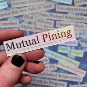May include: A rectangular sticker with the words "Mutual Pining" in a dark font. The sticker is held by a hand with black nail polish. Other small stickers with text are in the background.