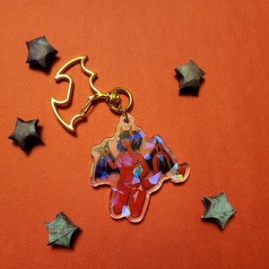 May include: A red and black demon character with horns and wings is featured on a clear acrylic keychain. The keychain is attached to a gold bat-shaped charm with a metal ring.