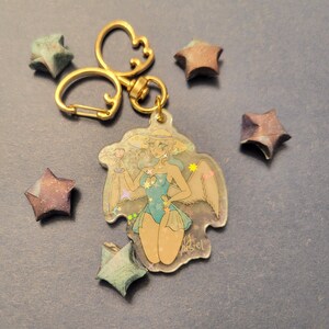 May include: A gold key chain with a clear resin charm featuring a cartoon angel with blue wings and a blue swimsuit. The charm is surrounded by iridescent glitter and has a light blue background. The charm is attached to a gold key ring with a split ring.