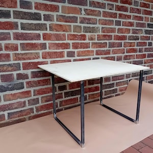 Könnte beinhalten: Eine weiße Tischplatte mit einem schwarzen Metallrahmen. Der Tisch steht auf einer braunen Oberfläche vor einer Backsteinwand.