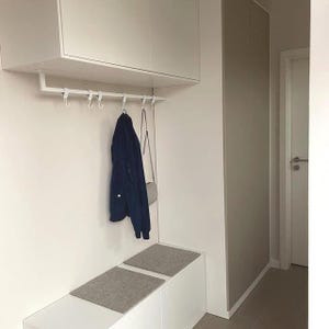 Könnte beinhalten: Ein moderner Eingangsbereich mit einer weißen Bank, Stauraum über Kopf und einer Garderobe. Eine marineblaue Jacke und eine hellgraue Handtasche hängen an der Garderobe. Zwei graue Filzmatten liegen auf der Bank.