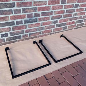 Könnte beinhalten: Zwei schwarze Metalltischbeine mit quadratischer Form und geschweißter Konstruktion. Die Beine stehen auf einer braunen Oberfläche.