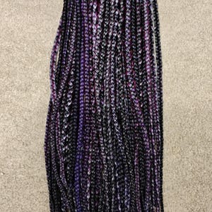 May include: A bundle of long, braided hair extensions in shades of black, purple, and white. The braids are tightly woven and hang down, creating a full and textured look. The hair is styled in a neat and organized manner.