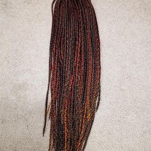 May include: A bundle of long, thin synthetic hair braids featuring a multi-tonal color palette of deep black, dark brown, and vibrant copper orange. The individual braids are tightly woven and hang vertically in a uniform, straight arrangement.
