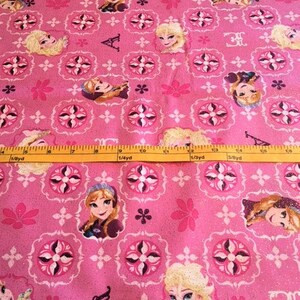 May include: Pink fabric with a repeating pattern of white and black tile designs. The tile designs feature images of the characters from the movie Frozen. The fabric is glittery.